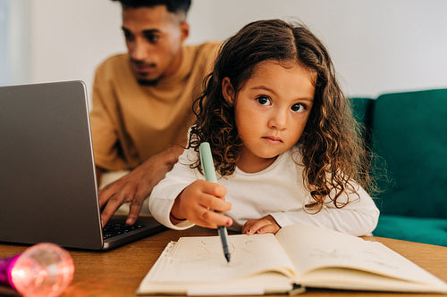 Little girl writing in a book while sitting with her father at home