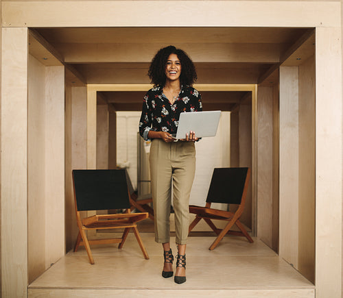 Portrait of a smiling woman entrepreneur standing in office holding laptop