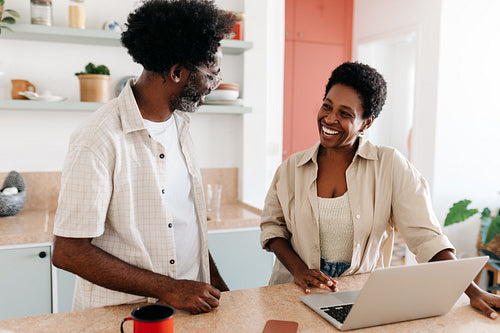 Mature black couple smiling, chatting and using a laptop together in the kitchen