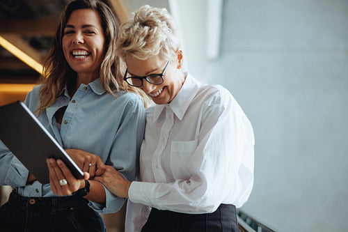 Two business women laugh happily as they use a digital tablet together in a modern office