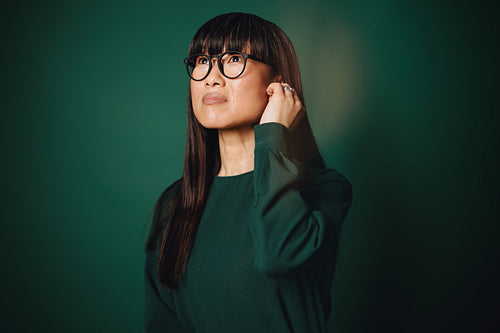 Thoughtful woman with glasses and earbuds looks up in a green portrait