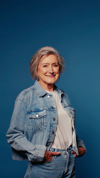Cheerful senior woman in denim jacket smiles for a studio portrait against a blue background