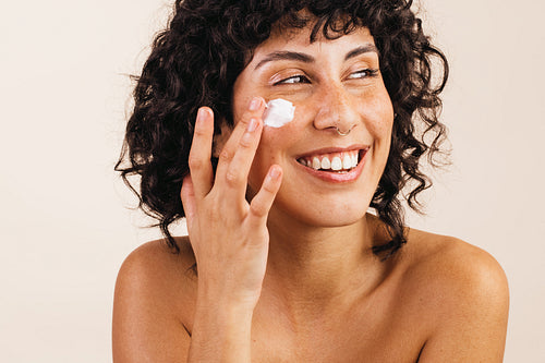 Happy young woman applying moisturizing cream on her face