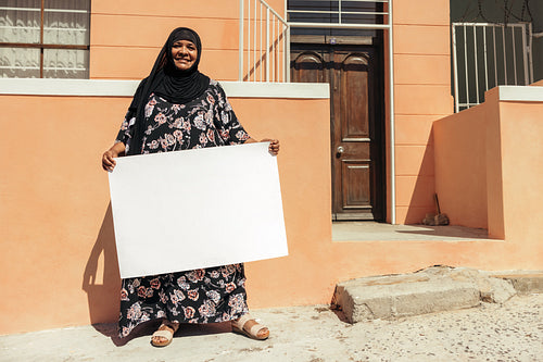 Smiling Muslim woman holding a blank placard outside her home