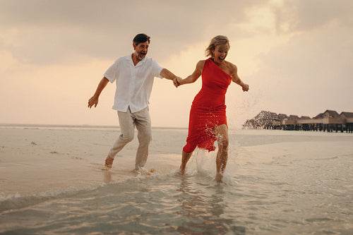 Playful mature couple enjoying a sunset at the island beach, with woman in red gown splashing in the water