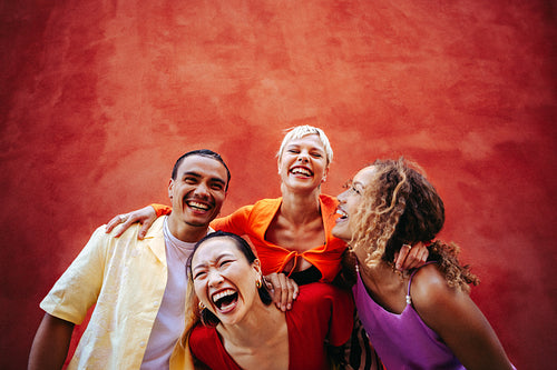 Happy friends laugh outdoors beside a red wall with their group standing close