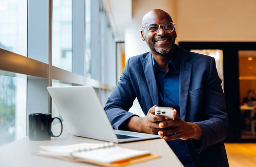 Smiling businessman using smartphone at a desk with laptop and notebook