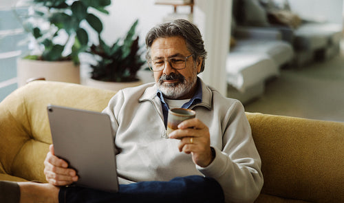 Seated senior man uses a tablet with coffee at home