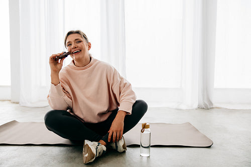 Eating a protein bar, the woman rests on her yoga mat after workout recovery