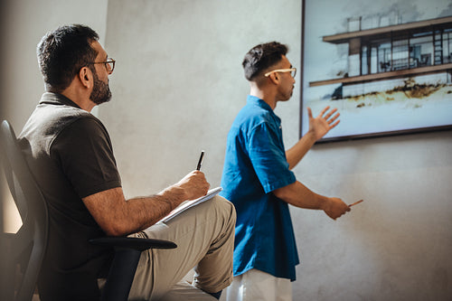 Middle-aged architect observing young colleague presenting home design concept in office.