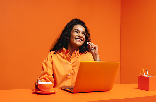 Gen z woman in an orange monochromatic setting using a laptop and holding drink, smiling and looking up