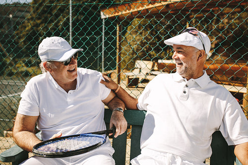 Two senior men sitting near a tennis court and talking
