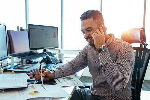 Entrepreneur discussing work over mobile phone sitting at his office desk.