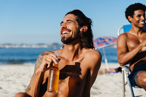 Young man enjoying a beer and laughing with friends at the beach