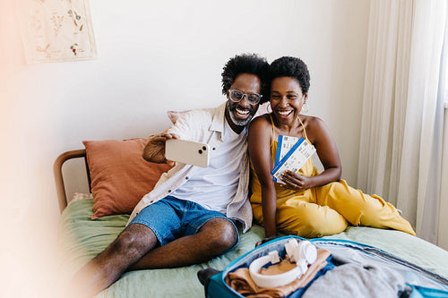 Excited couple smiling on bed with flight tickets, making a video call before a romantic getaway