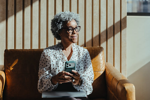 Mature woman using smartphone on sofa in modern office lounge