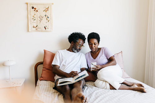 Relaxed mature couple enjoying quality time together in bed, reading and browsing on a mobile phone