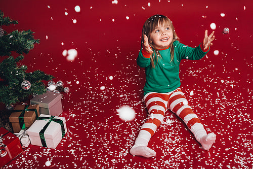Kid sitting beside a christmas tree enjoying artificial snowfall