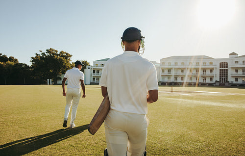 Two male cricketers walking on a sunny field with equipment