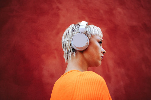 Blonde woman wears headphones for streaming outdoors by a red wall