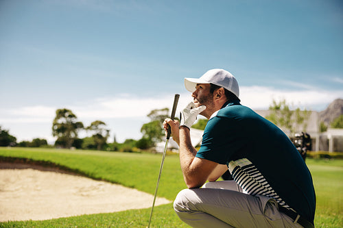 Focused male golfer crouching with a golf club near a sand bunker, strategizing his next move