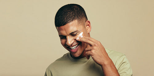 Happy with his skincare routine, young man smiles as he applies facial cream