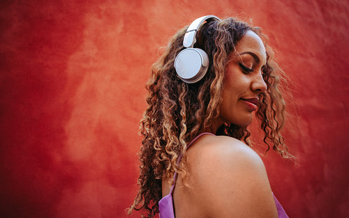 Woman immersed in music with headphones against red wall outdoors