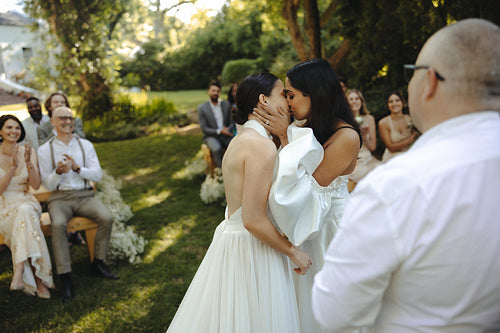 In love lesbian brides sharing a wedding kiss surrounded by guests in a lush garden