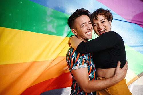 Smiling queer couple standing against a rainbow flag
