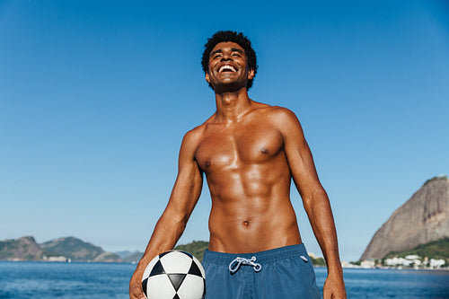 Shirtless young man with toned physique at the beach holding a soccer ball