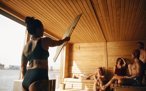 A wellness sauna session unfolds as a sauna master fans heat beside relaxing people.