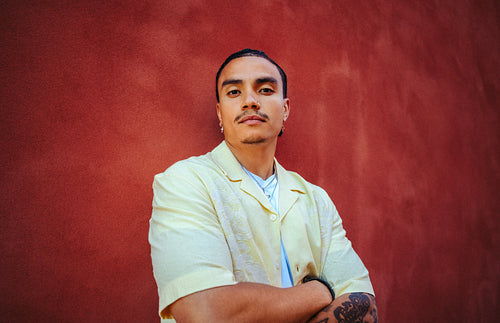 Latina man folds his arms in a portrait by a red wall outdoors