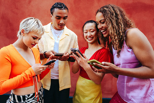 Multiracial group of friends shares social media content together outdoors against a bright wall