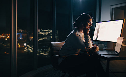 Female project manager working late in her home office, using a laptop and computer at her desk