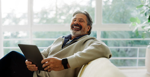Laughing senior man enjoys a tablet on the sofa