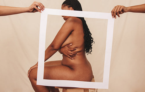 Naked woman sitting behind a picture frame in a studio
