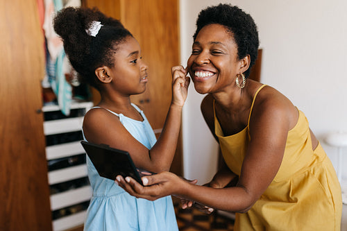 Mother smiling together as daughter applies makeup on her face in a bedroom
