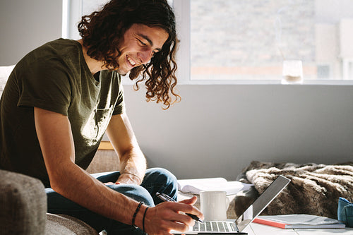 Smiling man studying at home on laptop with a pen in hand