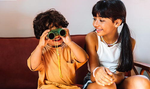 Two Latin American children sitting together playing with green binoculars and smiling