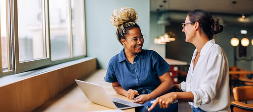 Two young women smiling and talking while working together at a laptop