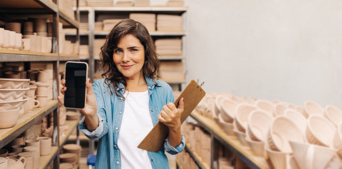 Confident ceramist showing a blank smartphone screen in her shop
