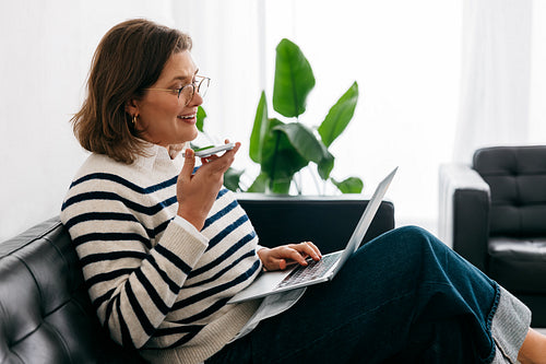 Speaking on her smartphone, the woman works from home on a couch with a laptop