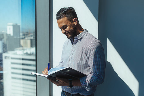 Business investor writing in diary standing at the window in office.