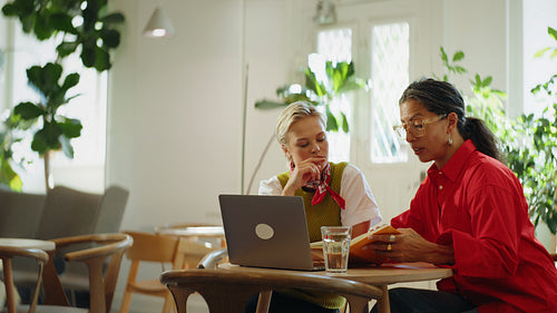 Women review a notebook beside a laptop during coworking at a cafe table
