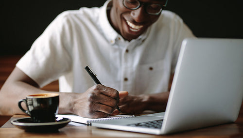 Man writing notes sitting at a coffee shop with a laptop on the table