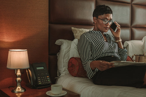 Businesswoman reading newspaper and talking on cell phone in hotel room