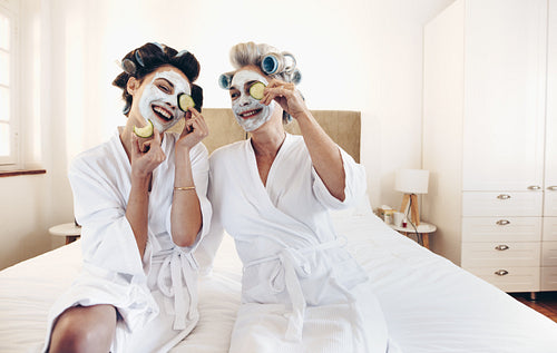 Happy women wearing beauty face pack sitting on bed holding slices of cucumber