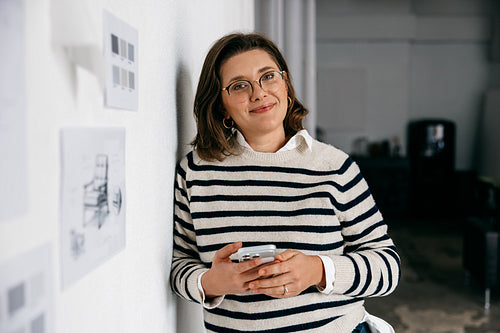 In a creative office, the woman checks her smartphone beside design sketches on the wall