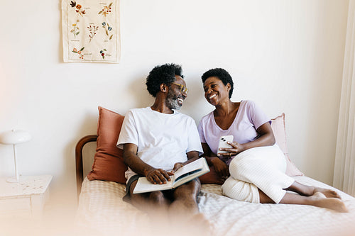 Happy mature couple enjoying relaxation time together in bed, reading and browsing on a smartphone