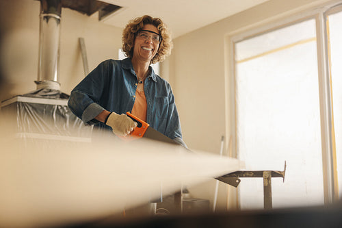 Happy mature woman in safety goggles working on a home renovation project with a crosscut saw